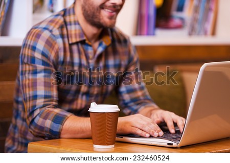 Working with pleasure. Close-up of young man working on laptop and smiling while sitting at the desk with bookshelf in the background