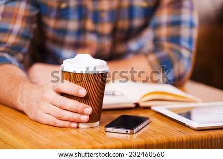 Taking time for coffee break. Close-up of man holding coffee cup while sitting at the desk with note pad and digital tablet laying on it