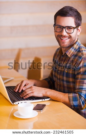 Having opportunity to work everywhere. Handsome young man working on laptop and smiling while sitting in cafe