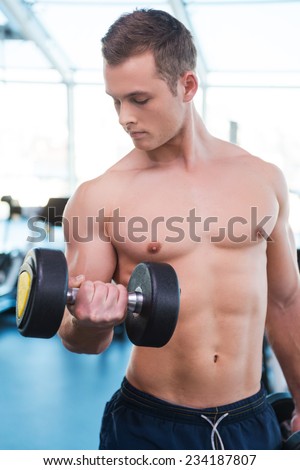 Training his muscles. Confident young muscular man training with dumbbell while standing in gym