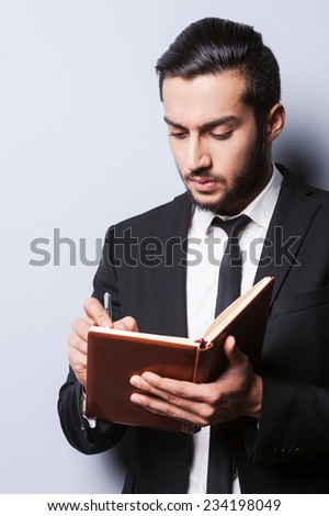I need some fresh ideas. Concentrated young man in formalwear holding note pad and writing in it with pen while standing against grey background
