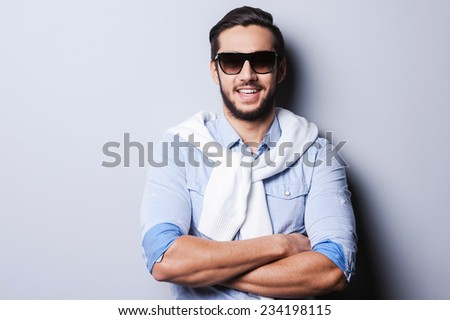 Charming handsome. Handsome young man in blue shirt keeping arms crossed and smiling at camera while standing against grey background
