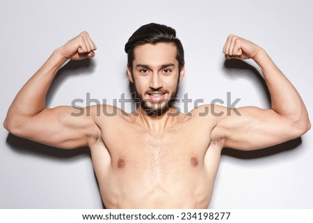 Strong man. Handsome young shirtless man keeping arms raised and looking at camera  while standing against grey background