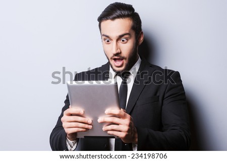 Examining his brand new tablet. Surprised young man in formalwear holding digital tablet while standing against grey background