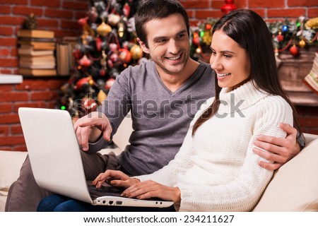 Just look at that! Beautiful young couple using computer together while man pointing monitor and smiling with Christmas decoration in the background