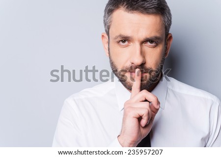 Keep my secret! Confident mature man in shirt and tie holding finger on lips and looking at camera while standing against grey background
