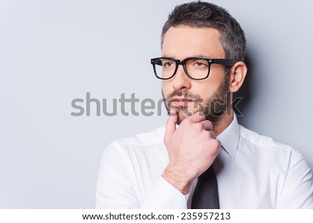 Lost in business thoughts. Portrait of thoughtful mature man in shirt and tie holding hand on chin and looking away while standing against grey background