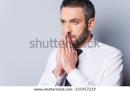 Thinking about solution. Portrait of concentrated mature man in shirt and tie holding hands clasped near face and looking away while standing against grey background