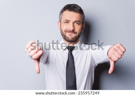 Bad news for you! Mature man in shirt and tie showing his thumbs down while standing against grey background