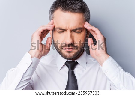 Feeling awful headache. Frustrated mature man in shirt and tie touching his head with fingers and keeping eyes closed while standing against grey background