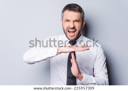 Time out! Furious mature man in shirt and tie shouting and gesturing time out while standing against grey background