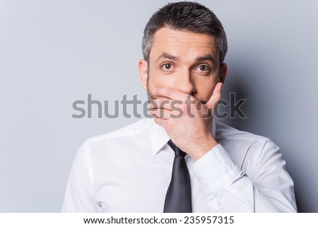 I am shocked! Surprised mature man in shirt and tie covering mouth with hand and looking at camera while standing against grey background