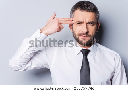 Game over! Frustrated mature man in shirt and tie gesturing handgun near head and looking at camera while standing against grey background