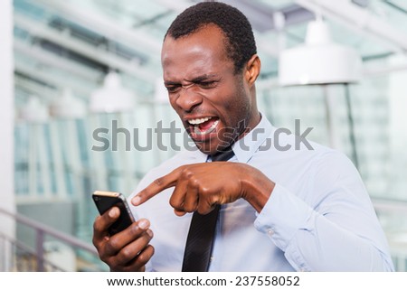 Bad news. Frustrated young African man in shirt and tie talking on the mobile phone and touching head with hand while standing indoors