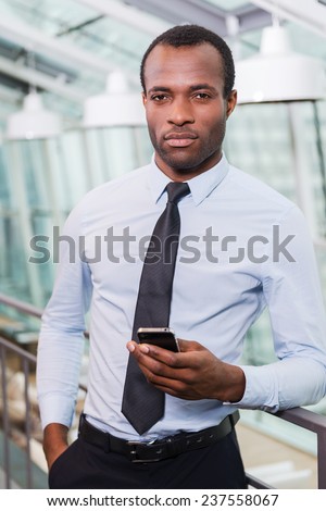 Confident businessman. Handsome young African man in shirt and tie holding mobile phone and looking at camera while standing indoors
