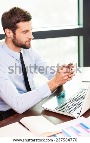 Typing business message. Top view of young man in shirt and tie holding mobile phone and looking at it while sitting at his working place
