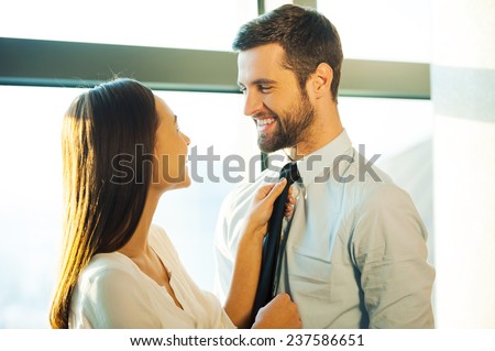 Now you are ready! Beautiful young loving couple standing face to face and smiling while woman adjusting necktie of her husband