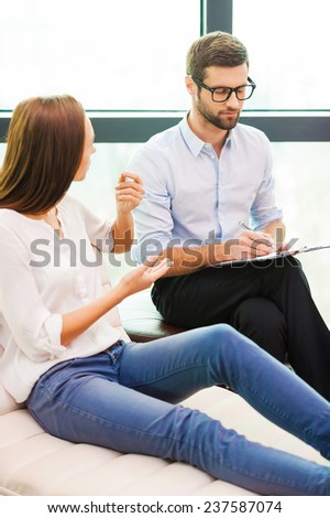 Sharing problems with professional. Worried young woman sitting at the chair and gesturing while male psychiatrist sitting close to her and writing something in his clipboard