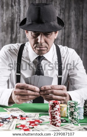Confident poker player. Close-up of serious senior man in shirt and suspenders sitting at the poker table and holding cards  with money and  gambling chips laying all around him