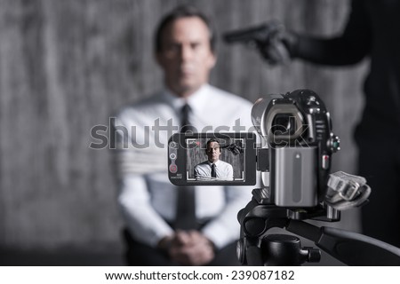 Filming hostage. Tied up businessman caught by a criminal sitting in front of a dirty wall with gun near his head while video camera filming it on the foreground