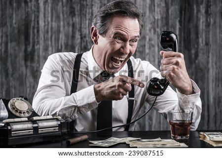 I will find you wherever you are! Furious senior man in shirt and suspenders shouting at telephone while sitting at the table with lots of money laying near him