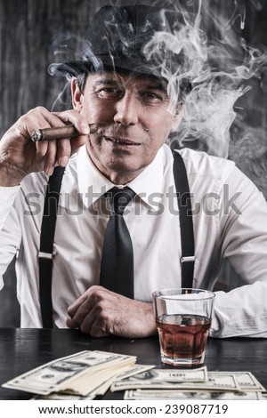 Mafia is not sleeping. Serious senior man in shirt and suspenders sitting at the table and smoking cigar while lots of money laying near him