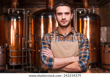 Confident brewer. Confident young male brewer in apron keeping arms crossed and looking at camera while standing in front of metal containers