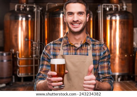 Happy brewer. Happy young male brewer in apron holding glass with beer and looking at it with smile while standing in front of metal containers
