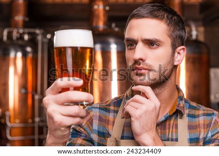 Controlling beer quality. Thoughtful young male brewer in apron holding glass with beer and looking at it while standing in front of metal containers