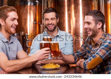 Meeting old friends in bar. Three happy young men in casual wear toasting with beer and smiling while sitting in beer pub together