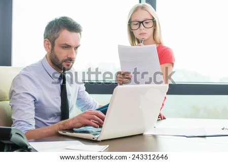 I want to be as my father. Confident young man in shirt and tie working on laptop while his little daughter sitting close to him and examining documents