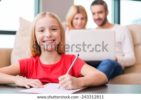 I love my family! Happy little girl drawing something on paper and smiling while her parents sitting in the background with laptop