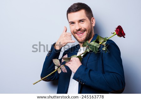 I will call you. Handsome young man carrying red rose on shoulders and gesturing mobile phone near face while standing against grey background