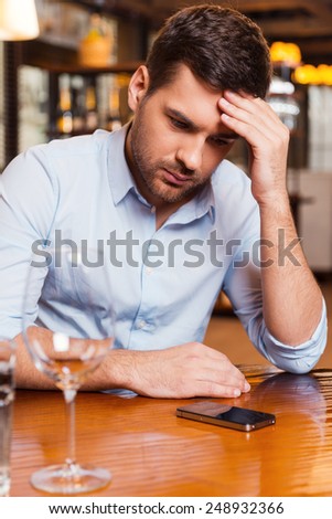 Waiting for her call. Frustrated young man holding head in hand and looking at the mobile phone while sitting at the restaurant