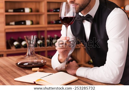 Sommelier examining wine. Cropped image of confident male sommelier examining wine while smelling it and leaning at the wooden table with wine shelf in the background