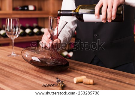 Sommelier at work. Confident male sommelier pouring wine to decanter while standing near the wine shelf