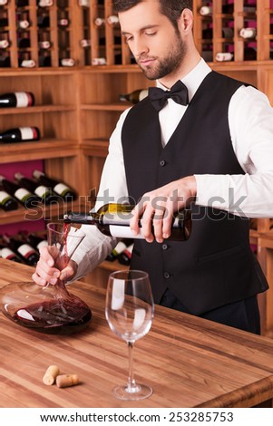 Pouring wine to decanter. Confident male sommelier pouring wine to decanter while standing near the wine shelf