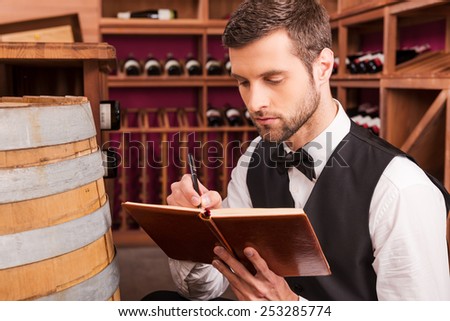 Making notes about wine. Confident male sommelier writing something in his note pad while sitting near the wine barrel