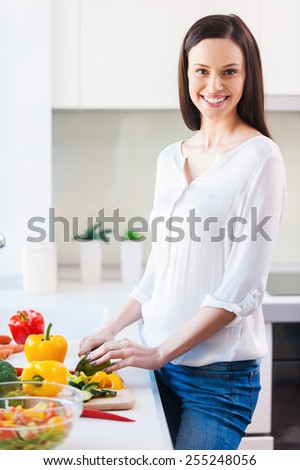 Using the freshest veggies for my meal. Beautiful young woman cutting vegetables and smiling while standing in the kitchen