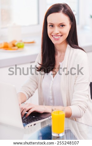 Surfing the net at home. Attractive young woman working on laptop and smiling while sitting in the kitchen