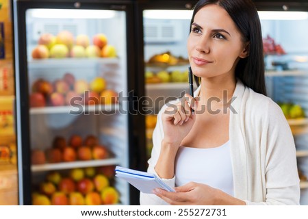 Woman with shopping list. Thoughtful young woman holding shopping list and looking away while standing in front of refrigerators in grocery store