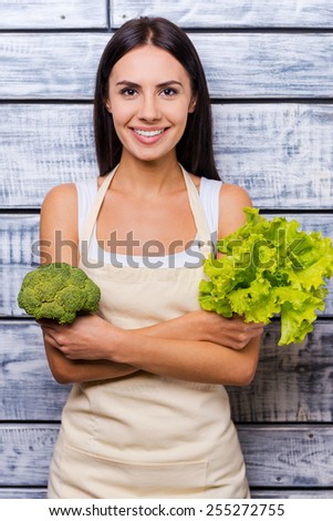 Eat healthy! Beautiful young smiling woman in apron holding fresh lettuce and broccoli while standing in front of wooden background