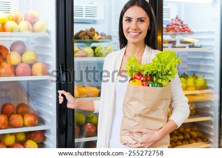 Buying the freshest products. Beautiful young woman holding shopping bag with fruits and smiling while standing in grocery store near refrigerator