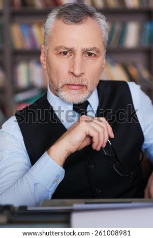 Confident author. Confident grey hair senior man in formalwear sitting at the typewriter and looking at camera with bookshelf in the background