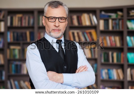 Confident and intelligence. Confident grey hair senior man in formalwear keeping arms crossed and looking at camera while standing against bookshelf