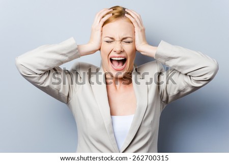 Everything goes wrong! Beautiful young businesswoman screaming and holding head in hands while standing against grey background