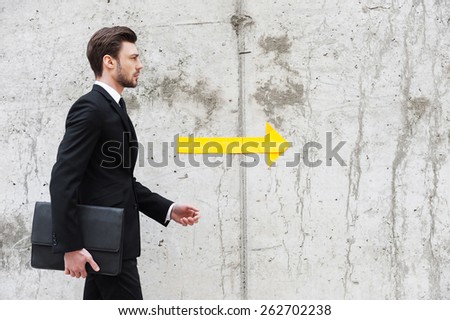 Right direction. Handsome young man in formalwear holding briefcase while walking in front of the concrete wall