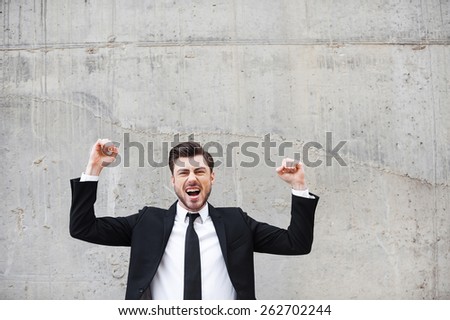 Everyday winner. Happy young man in formalwear keeping arms raised and expressing positivity while standing against the concrete wall