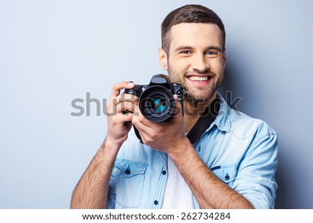 Give me a smile! Handsome young man holding digital camera and smiling while standing against grey background