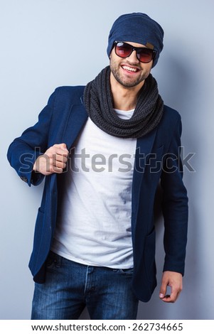 Confident in his perfect style. Handsome young stylish man in sunglasses and hat adjusting his jacket and looking at camera while standing against grey background